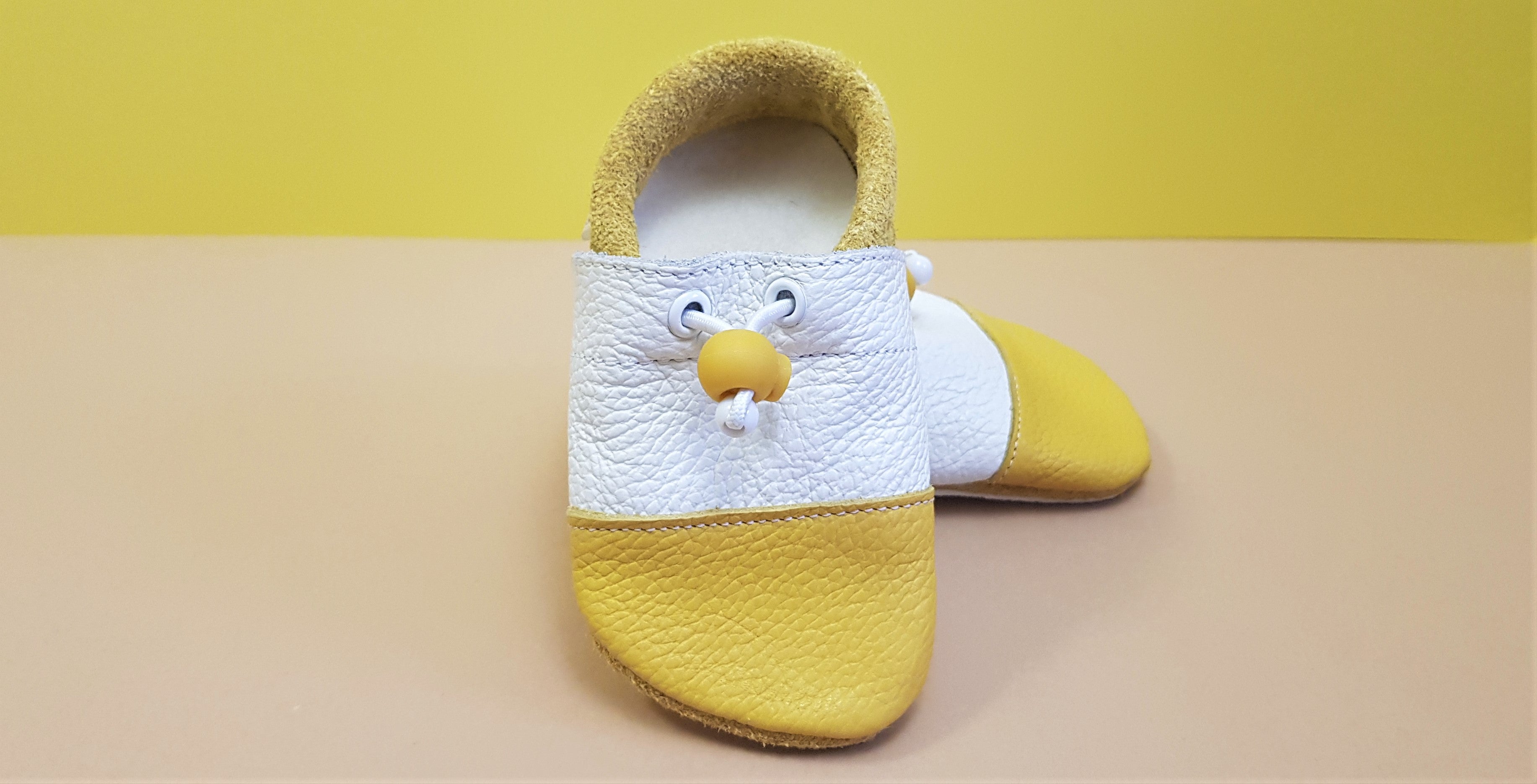 Yellow and white Nohatka soft sole booties/slippers for babies and toddler, anatomically shaped from soft leather with adjustable ankle fastening and anti slip indoor/outdoor sole
